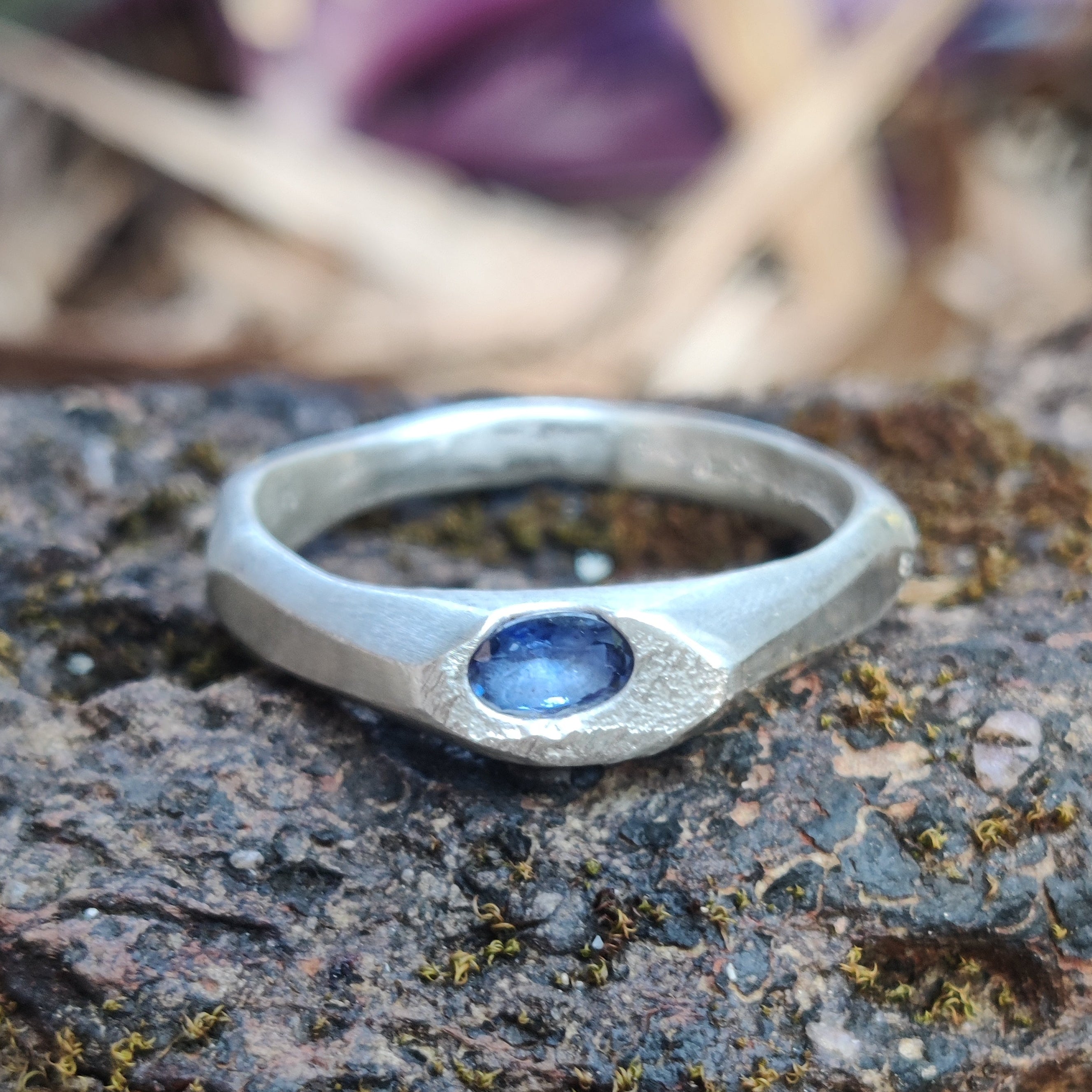 Silver ring with a blue stone on a textured surface with blurred purple flowers in the background