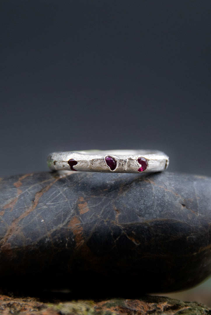 Three Little Ruby sandcasting ring featuring unique textures and three embedded ruby stones resting on a stone surface.
