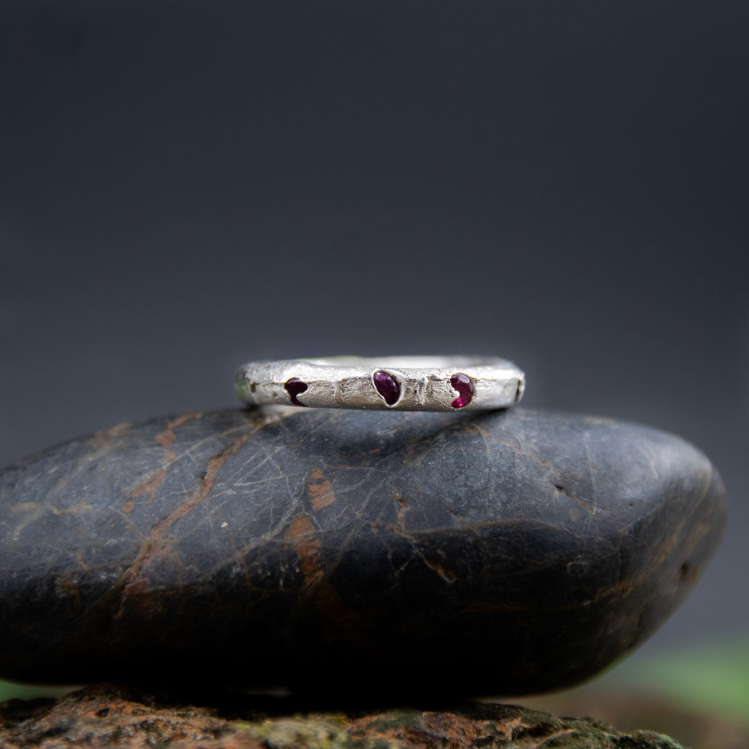 Three Little Ruby sandcasting ring featuring unique textures and three embedded ruby stones resting on a stone surface.